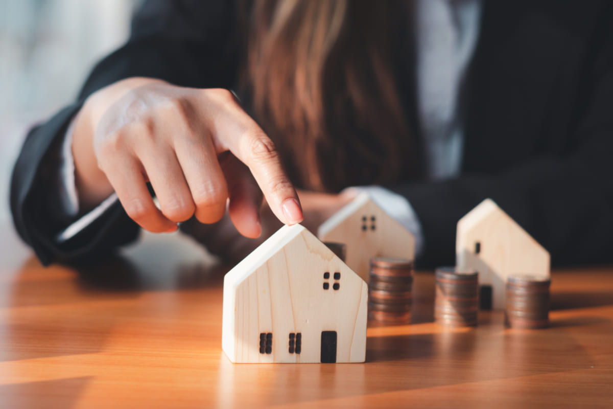 A woman in a suit touching a wooden model house next to coins