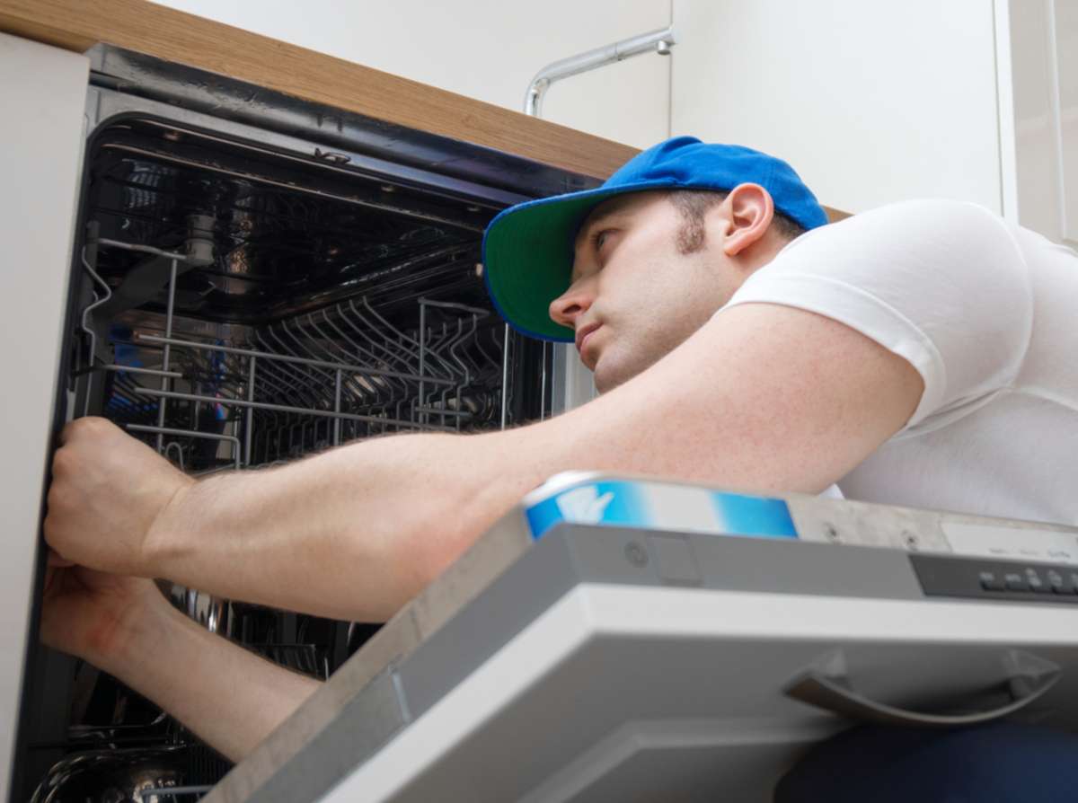 Professional handyman in overalls repairing domestic dishwasher in the kitchen