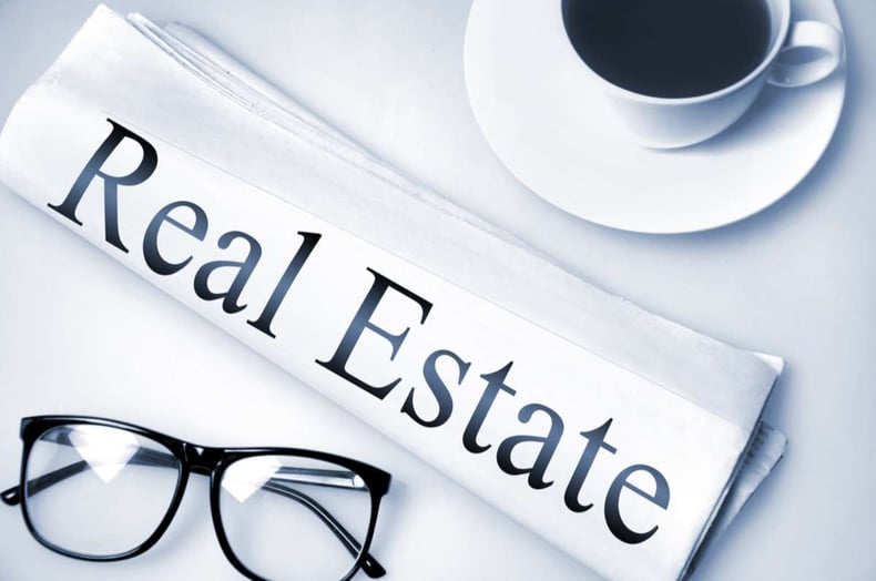 The words real estate on a newspaper next to glasses and a coffee