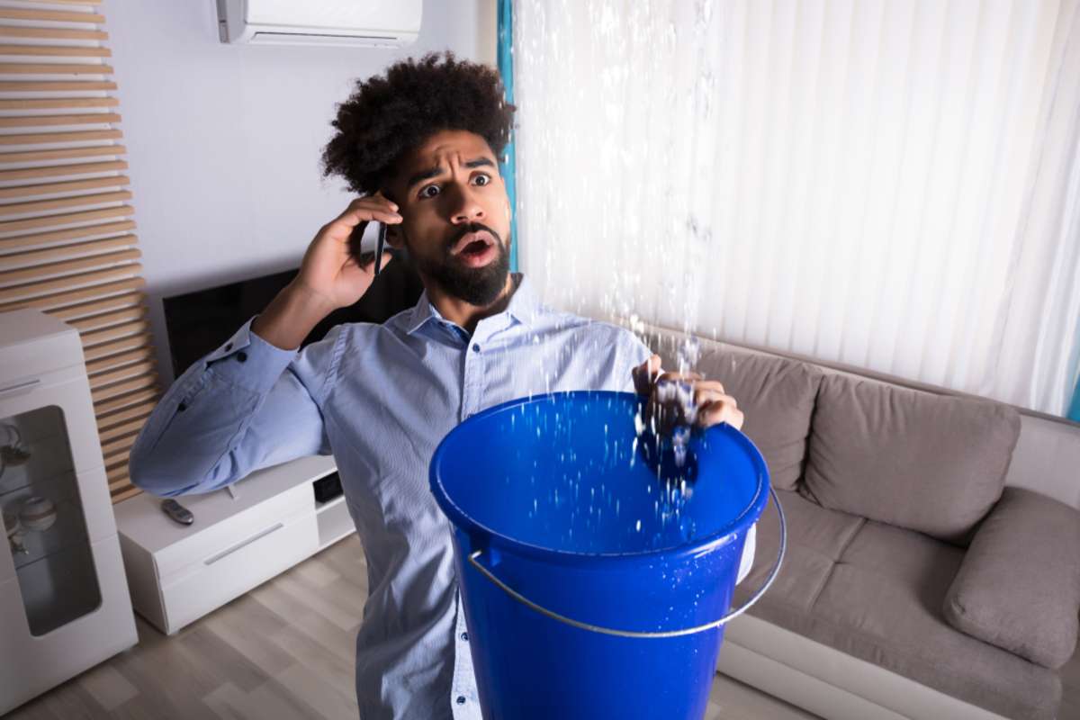 Man on the phone while water falls from the ceiling into a bucket, emergency maintenance concept. 