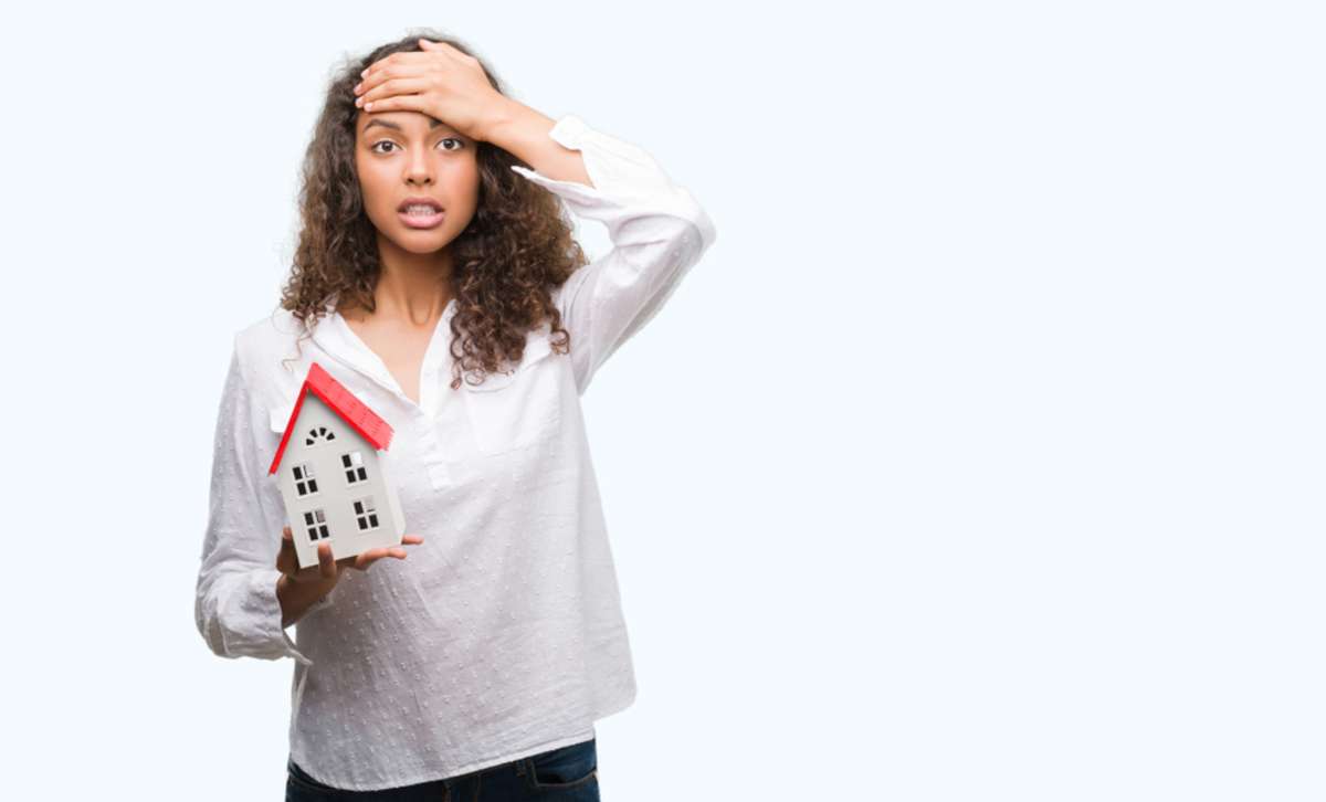 Young woman holding small house stressed with hand on head, not having Hampton property management concept. 