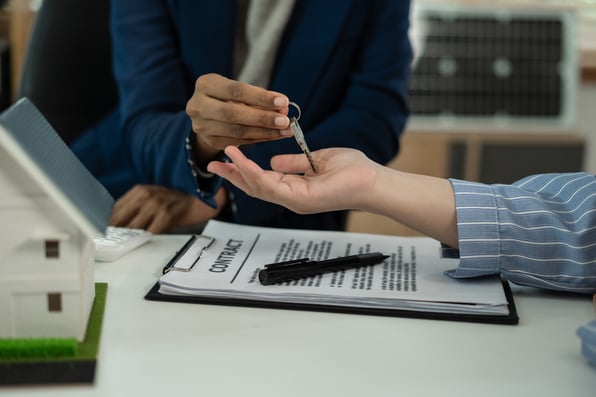 A man in a suit handing a woman keys next to a lease agreement and a model home.